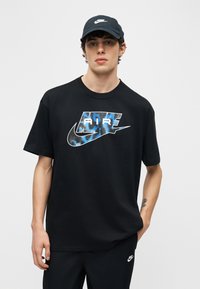 Bărbat tânăr purtând o șapcă neagră Nike, tricou negru cu logo albastru și alb Nike Air și pantaloni negri Nike, stând în fața unui fundal simplu.