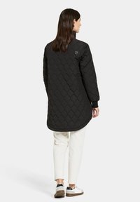 Svart quiltad lång jacka med hög krage, prydd med ett diamantmönster och en slimmad design. Kombinerad med cremefärgade byxor och vita sneakers.