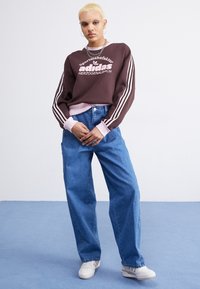 Brun sweatshirt med vit Adidas-logotyp, rosa detaljer och randiga ärmar. Matchad med ljusblå vida jeans och vita sneakers.