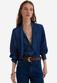 Donkerblauwe button-up overhemd met een gedrapeerde kraag, gecombineerd met high-waisted donkere jeans en een getextureerde bruine riem met een goudkleurige gesp.