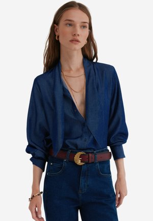 Donkerblauwe button-up overhemd met een gedrapeerde kraag, gecombineerd met high-waisted donkere jeans en een getextureerde bruine riem met een goudkleurige gesp.