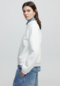 Sudadera blanca de corte holgado y puños acanalados, superpuesta a una camisa de denim. Modelo de pie contra un fondo claro y liso.