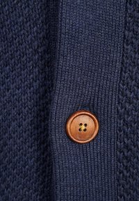 Mörkblå stickad kofta med ett texturerat mönster, med en träknapp med fyra hål som framhäver dess hantverk och detaljrikedom.