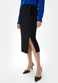Zwarte wikkelrok met een split aan de voorkant en een zijstrikdetail, gedragen met een blauwe gebreide trui en zwarte hoge hakken. Glad textuur.