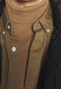 Un col roulé marron à côtes superposé sous une chemise boutonnée beige clair avec des boutons en perles, accompagné d'une veste sombre sur le côté.