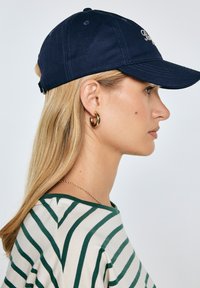 Žena s dlouhými blond vlasy, která má na sobě námořnickou čepici, zlaté kruhové náušnice, zeleno-bíle pruhované tričko a jemný zlatý řetízek, otočená doprava.