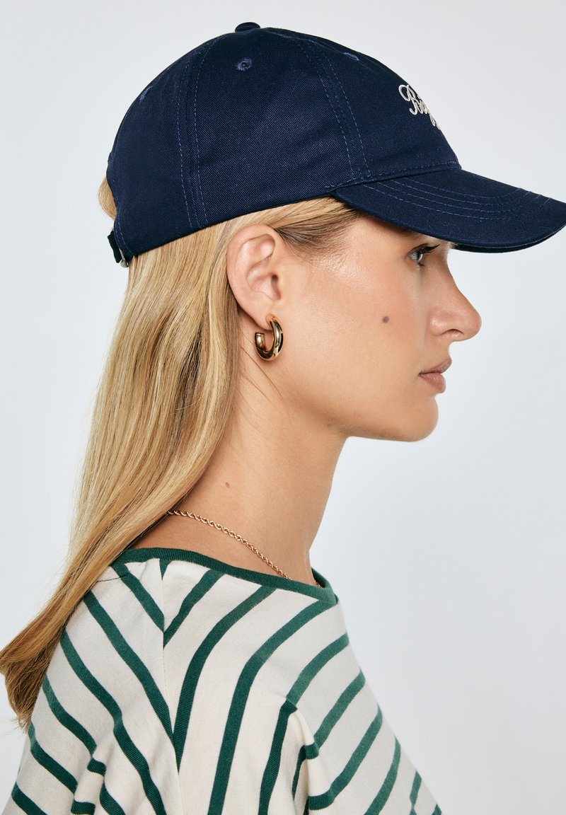 Žena s dlouhými blond vlasy, která má na sobě námořnickou čepici, zlaté kruhové náušnice, zeleno-bíle pruhované tričko a jemný zlatý řetízek, otočená doprava.
