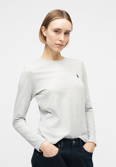 Γυναίκα με κοντά ξανθά μαλλιά, φορώντας ανοιχτό γκρι μακρυμάνικο πουκάμισο με ένα μικρό μπλε κεντητό λογότυπο και σκούρο παντελόνι, χέρια στις τσέπες.