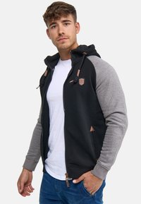 Sudadera con cremallera en negro y gris, con capucha ajustable, un parche con logo y mangas en contraste. Presenta un tejido texturizado y detalles en hardware de bronce.