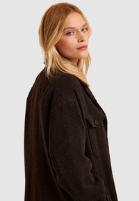 Chaqueta de gran tamaño en marrón oscuro, hecha de tejido texturizado, con cuello y un bolsillo delantero con cierre de botón.