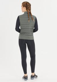 Vadderad grå väst med hög krage, bärs över långa svarta ärmar, i kombination med svarta leggins och sportskor. Minimalistisk design.