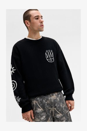 Musta fleece-pusero, jossa on valkoiset graafiset kuvastot hihoissa ja rinnassa, pyöreä kaula-aukko ja rento malli, yhdistettynä kuviollisiin housuihin.