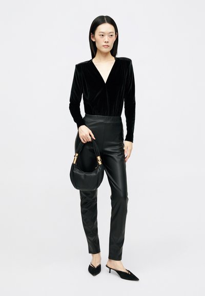 Top lung din catifea neagră cu mâneci lungi și decolteu adânc în V, asociat cu pantaloni strâmți din piele sintetică neagră. Modelul ține o geantă neagră cu accente aurii.