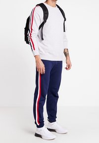 Sudadera gris con rayas rojas y azules en las mangas, combinada con pantalones deportivos azul marino con rayas laterales, y zapatillas blancas. Lleva una mochila negra.