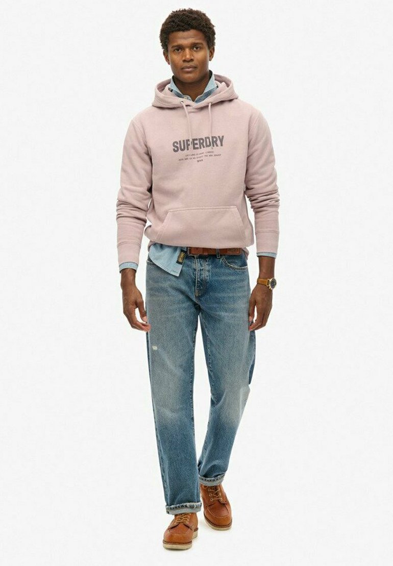 Lichtroze hoodie met "SUPERDRY" logo, gelaagd over een denim shirt. Draag het met donkere jeans en bruine veterschoenen. Casual outfitontwerp.
