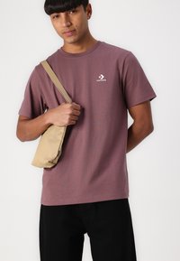 Kratek rokav, mauve T-shirt z okroglim ovratnikom in majhnim belim logotipom Converse na prsih, v kombinaciji s bež torbico preko telesa.