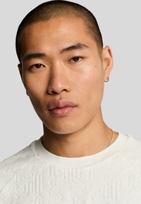 Vit texturerad tröja med rundad halsringning. Slät hy med kortklippt frisyr, bär små hoops av silver.