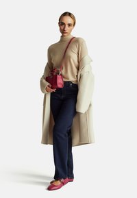 Beżowy sweter z golfem, kremowy płaszcz, ciemnoniebieskie dżinsy, różowa torebka na krótkich uchwytach oraz różowe płaskie buty z klamrą.