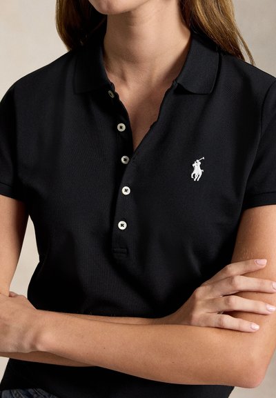 Γυναίκα με κοντομάνικο μαύρο πόλο πουκάμισο με λευκό λογότυπο αλόγου και αναβάτη, σταυρωμένα χέρια, μπεζ φόντο.