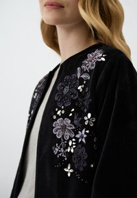 Cardigan nero con ricamo floreale in viola e bianco, con perline e paillettes, morbida texture, design aperto e senza chiusure.