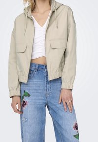Beige cropped jacket with a hood, side pockets, and elastic cuffs, paired with blue jeans featuring floral embroidery and a distressed knee.