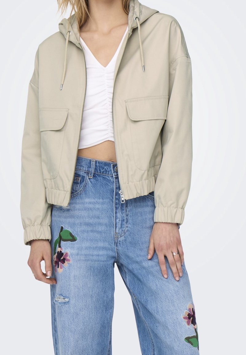 Beige cropped jacket with a hood, side pockets, and elastic cuffs, paired with blue jeans featuring floral embroidery and a distressed knee.