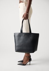 Svart skinn tote veske med flat design, to skulderhåndtak, gull logo på fronten, holdt av en person i beige bukser og svarte hæler.