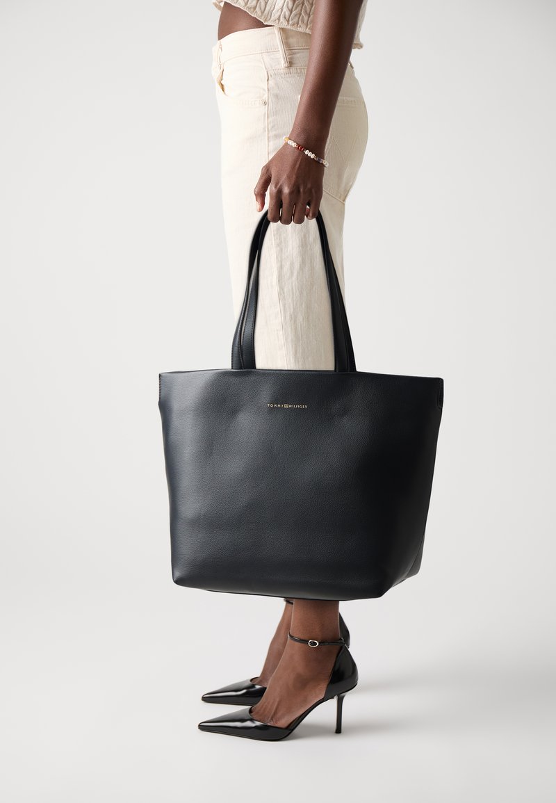 Svart skinn tote veske med flat design, to skulderhåndtak, gull logo på fronten, holdt av en person i beige bukser og svarte hæler.