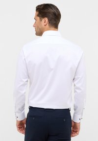 Weißes Langarmhemd mit strukturiertem Kragen und geknöpften Manschetten, kombiniert mit marineblauen Hosen, die über zwei Gesäßtaschen und Knöpfe verfügen.