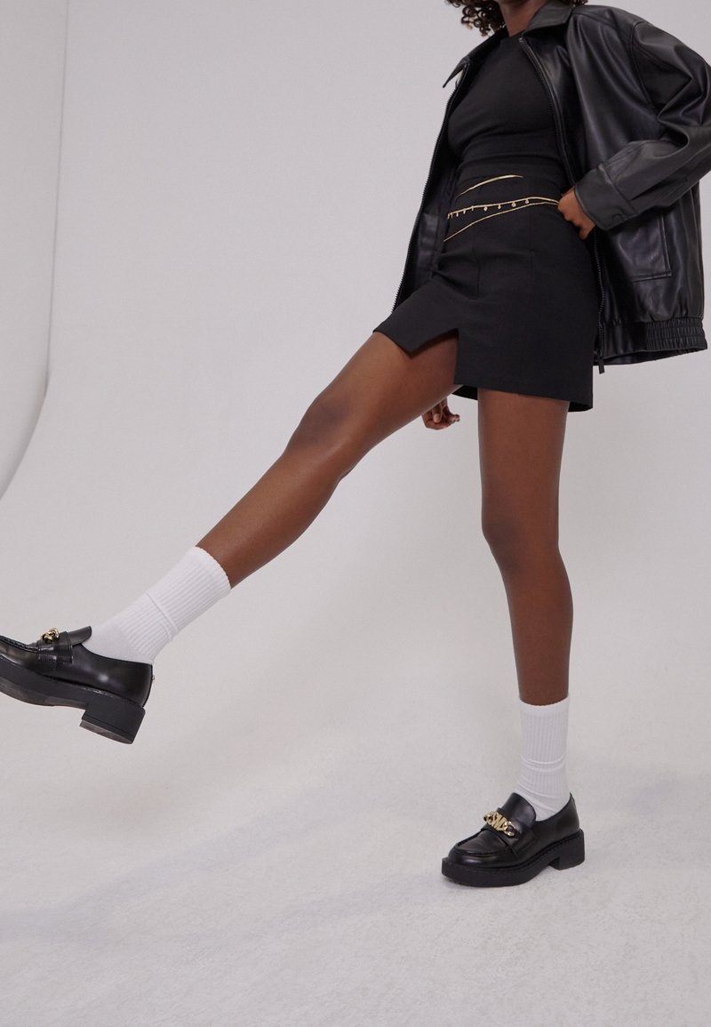 Personne portant une veste en cuir noire, une mini-jupe noire avec des chaînes dorées, des mocassins noirs avec des chaînes dorées et des chaussettes blanches, levant une jambe vers l'avant.