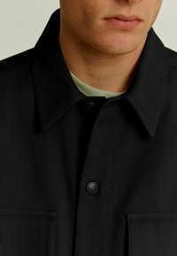 Zwarte overhemdjas met een kraag, knoopsluiting en een gestructureerde stof met zichtbare stiksels en een borstzak. Lichtgroen ondershirt.