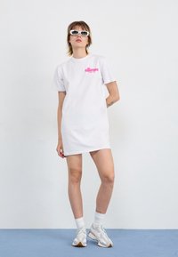 Jeune personne debout portant une robe t-shirt blanche avec un logo rose, des baskets blanches, des chaussettes, des lunettes de soleil rectangulaires blanches et des boucles d'oreilles anneaux sur un sol bleu.