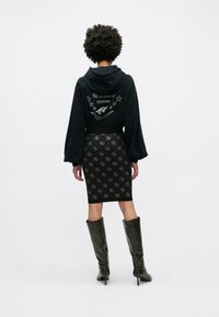 Schwarzer Hoodie mit grafischem Design auf der Rückseite, kombiniert mit einem schwarzen, gemusterten Bleistiftrock. Schwarze Stiefel mit Absatz vervollständigen das Outfit.