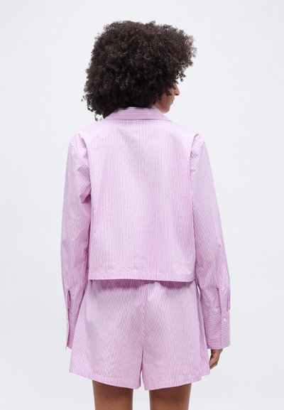 Γυναίκα με σγουρά μαλλιά, που φοράει πουκάμισο μακρυμάνικο με κάθετες ριγέ ροζ και λευκές λεπτομέρειες και σορτς σετ, με θέα από πίσω.