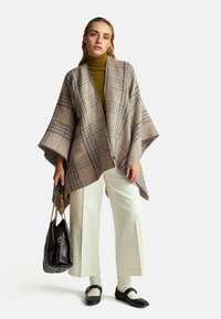 Geruite poncho in beige met marineblauwe en bruine accenten, gecombineerd met een mosterdgele coltrui, crèmekleurige wijde broek en een zwarte handtas.