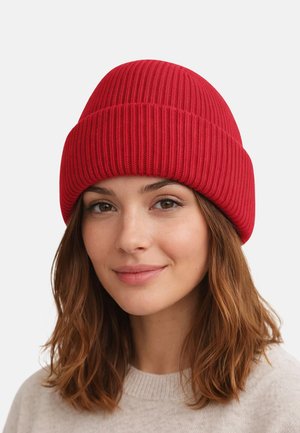 Young woman with shoulder-length brown hair wearing a bright red ribbed knit beanie and a light beige sweater, smiling softly.