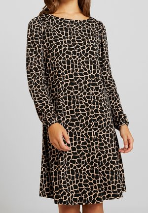 Kobieta ubrana w sukienkę z długim rękawem sięgającą kolan, z beżowo-czarnym nieregularnym geometrycznym wzorem na jasnoszarym tle.