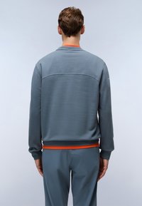 Grå hettegenser med teksturert overflate, rund hals og oransje aksent ved kanten. Designet for en avslappet passform, vist fra modellens bakside.