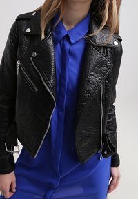 Black leather biker jacket with multiple zippers and silver hardware, layered over a blue shirt. The texture is slightly crinkled.