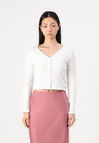Cardigan blanc à côtes avec manches longues, décolleté en V et fermeture à boutons, porté sur une jupe en satin rose. Coupe courte avec une texture lisse.