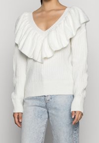 Maglione bianco con scollatura a V profonda, caratterizzato da un ampio colletto arruffato e una texture a costine. Abbinato a jeans azzurri chiari.