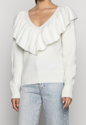 Suéter blanco con un escote en V profundo, que presenta un gran collar con volantes y textura acanalada. Combinado con jeans de color azul claro.
