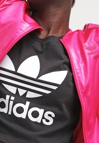 Fuchsiasattin zip-jacka med vit Adidas-logga på högra axeln, bärs över en svart t-shirt med en stor vit trefoil-logga.