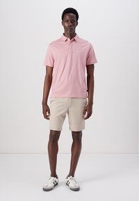 Pink short-sleeve polo shirt with a collar, paired with beige shorts. Model wears black and white sneakers. Simple, casual design.
