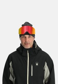 Skibrille mit einer großen, gebogenen rot-goldenen Spiegellinse, getragen über einem schwarzen Hut und einer schwarzen Jacke mit grauen Akzenten und Logo.