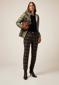 Olivengrøn puffjakke, marineblå sweater, brune ternede bukser, sorte ankelstøvler. Holder en brun håndtaske med guldkædedetalje.