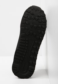Semelle noire de chaussure avec des crampons profonds et "VERSACE JEANS" embossé sur la zone de la voûte centrale, incliné sur un fond blanc.