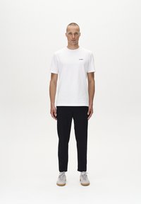 Camiseta blanca de algodón con mangas cortas, que presenta un logo negro en el pecho. Combinada con pantalones negros ajustados y zapatillas grises.