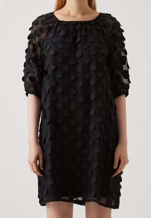 Frau trägt ein schwarzes durchsichtiges Kleid mit großen strukturierten Polka-Punkten, Dreiviertelärmeln und rundem Ausschnitt.