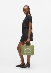 Μαύρο τοπ κοντό μανίκι σε συνδυασμό με μαύρες σορτς, κρατώντας μια πράσινη τσάντα Karl Lagerfeld με λευκό κείμενο, φορεμένο με μαύρες επίπεδες παπούτσια με τρουκς.